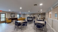 This well-appointed single-floor community space offers a thoughtfully designed layout that seamlessly blends dining, socializing, and recreation. The expansive main lounge is filled with recessed lighting and features light wood-tone tile flooring throughout, creating a bright and welcoming atmosphere. Multiple dining tables paired with navy blue upholstered chairs provide ample seating, while a raised bar-height counter area along one wall adds a casual gathering option.

A dedicated lounge nook offers a more intimate setting, furnished with mustard yellow accent chairs and a sleek electric fireplace set within a rich wood console. Framed artwork and views of the greenery outside through glass doors enhance the relaxed ambiance of this area.

The kitchen area is generously sized, equipped with dark cabinetry, granite countertops, a mobile butcher block island, a dishwasher, coffee stations, and a wall-mounted television — ideal for food prep and service. A pass-through window connects the kitchen to the dining area for added convenience.

Deeper into the floor plan, a recreation room houses a billiards table set atop dark decorative flooring, offering a distinct space for leisure activities. Connecting corridors with white wainscoting and recessed lighting lead throughout the facility, reinforcing the clean, institutional-yet-comfortable design aesthetic of the overall space.