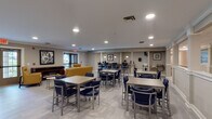 This well-appointed single-floor community space offers a thoughtfully designed layout that seamlessly blends dining, socializing, and recreation. The expansive main lounge is filled with recessed lighting and features light wood-tone tile flooring throughout, creating a bright and welcoming atmosphere. Multiple dining tables paired with navy blue upholstered chairs provide ample seating, while a raised bar-height counter area along one wall adds a casual gathering option.

A dedicated lounge nook offers a more intimate setting, furnished with mustard yellow accent chairs and a sleek electric fireplace set within a rich wood console. Framed artwork and views of the greenery outside through glass doors enhance the relaxed ambiance of this area.

The kitchen area is generously sized, equipped with dark cabinetry, granite countertops, a mobile butcher block island, a dishwasher, coffee stations, and a wall-mounted television — ideal for food prep and service. A pass-through window connects the kitchen to the dining area for added convenience.

Deeper into the floor plan, a recreation room houses a billiards table set atop dark decorative flooring, offering a distinct space for leisure activities. Connecting corridors with white wainscoting and recessed lighting lead throughout the facility, reinforcing the clean, institutional-yet-comfortable design aesthetic of the overall space.