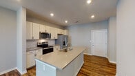 This well-appointed single-floor unit offers a clean, modern aesthetic throughout, combining functionality with contemporary design. The heart of the home is an open-concept kitchen that flows seamlessly into the living area. The kitchen is equipped with a full suite of stainless steel appliances, including a range, microwave, and French door refrigerator with an in-door water and ice dispenser. Crisp white cabinetry extends to the ceiling for generous storage, while a classic subway tile backsplash adds a timeless touch. A large center island with a quartz countertop and an undermount sink serves as both a practical workspace and a natural gathering spot. Rich, warm-toned hardwood-style flooring runs throughout the main living areas, adding depth and character to the open layout. The bedroom features plush carpeting, a ceiling fan with integrated lighting, and a window with natural light. It connects directly to a walk-in closet with built-in shelving and a private bathroom. The bathroom is finished with a wide quartz vanity, an undermount sink, modern fixtures, and a large mirror. The overall palette of soft gray walls and white trim creates a bright, neutral backdrop that feels both welcoming and move-in ready.