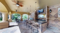 This covered outdoor entertaining space is designed with both comfort and style in mind. At its centerpiece is a spacious built-in bar with a dark countertop, accommodating several ornate wrought-iron barstools that add a classic, decorative touch. The bar area is equipped with a sink and backed by a striking tile wall featuring a diamond-pattern design in warm earth tones, accented by a decorative medallion motif. A wall-mounted flat-screen TV is positioned for easy viewing from the bar seating.

Overhead, a combination of ceiling fans and pendant lights ensures comfortable airflow and warm ambient lighting throughout the space. The flooring is composed of natural stone tile, contributing to the upscale, resort-like atmosphere. Elegant arched columns frame the open sides of the covered area, seamlessly connecting the interior to the surrounding outdoor environment, where mature trees and lush landscaping are visible. Additional seating is available beyond the bar area, offering flexible arrangements for gatherings. The overall design blends Mediterranean-inspired architectural details with practical outdoor living amenities.