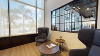 This contemporary single-floor office space is thoughtfully designed to balance professional functionality with an inviting atmosphere. The interior is characterized by soaring ceilings, crisp white walls, and large floor-to-ceiling windows that flood the space with natural light and frame views of outdoor palm trees, lending a distinctly warm, coastal character to the environment.

The reception and lounge area serves as a focal point upon entry, featuring a pair of high-backed upholstered wingback chairs arranged around a round wooden coffee table, accented by a small decorative globe and reading materials. An interior black-framed grid window connects this lounge visually to an adjacent workspace, maintaining an open, connected feel while defining distinct zones within the office.

Entry is made through bold black-framed glass double doors with transom windows above, reinforcing the space's modern industrial aesthetic. A natural wood slatted room divider provides a warm contrast to the sleek glass and black metal elements throughout.

A private meeting room features a round dark table surrounded by upholstered chairs and is distinguished by a dramatic full-wall vintage aircraft mural, adding a striking visual identity to the space. The workstation area beyond benefits from exposed ceiling details and track lighting, completing a polished, design-forward professional environment.
