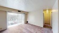 This single-floor unit offers a practical and functional layout across several well-defined spaces. The living room is the heart of the unit, featuring plush carpeting, a textured ceiling, and a large sliding glass door dressed with vertical blinds that floods the room with natural light and provides views of the surrounding exterior. The open feel of the living area is complemented by neutral wall tones that create a clean, inviting atmosphere.

The kitchen is compact yet efficient, showcasing rich dark cabinetry with ample upper and lower storage, a marble-patterned laminate countertop, a stainless steel sink, and a full-size refrigerator — offering everything needed for everyday cooking and meal preparation.

The bedroom is a comfortable, carpeted retreat with a window fitted with blinds that allows for natural light while maintaining privacy. The textured ceiling continues throughout, lending a consistent character to the space.

A secondary room features a striking accent brick wall and a ceiling fan with a light fixture, adding a touch of warmth and charm to what could serve as a den or additional living area.

The bathroom is clean and straightforward, featuring a full tub-shower combination with built-in shelving, a toilet, and a vanity with a mirrored medicine cabinet, all finished in crisp white tones.