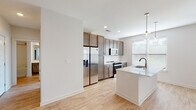 This well-designed home offers a thoughtful layout spread across multiple levels, connected by a carpeted staircase with a clean white railing. The open-concept kitchen serves as a central highlight, featuring rich medium-toned cabinetry that extends to the ceiling, a white quartz waterfall island with an integrated sink, stainless steel appliances including a French door refrigerator and electric range, a subway tile backsplash, and pendant glass globe lighting. Multiple windows invite abundant natural light throughout the kitchen and adjoining living area, which features light hardwood-style flooring, recessed lighting, and a ceiling fan. The living space opens toward a front entry door framed by greenery views. Bedrooms are generously sized, each equipped with ceiling fans, plush neutral carpet, and windows offering outdoor views. The primary bathroom is elongated and well-appointed, featuring a dual-sink vanity with dark cabinetry, white countertops, light wood-style flooring, and access to a walk-in closet. A secondary bathroom includes a soaking tub with a tiled surround and a single-sink vanity, maintaining the same cohesive design aesthetic found throughout the home.