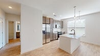 This well-designed home offers a thoughtful layout spread across multiple levels, connected by a carpeted staircase with a clean white railing. The open-concept kitchen serves as a central highlight, featuring rich medium-toned cabinetry that extends to the ceiling, a white quartz waterfall island with an integrated sink, stainless steel appliances including a French door refrigerator and electric range, a subway tile backsplash, and pendant glass globe lighting. Multiple windows invite abundant natural light throughout the kitchen and adjoining living area, which features light hardwood-style flooring, recessed lighting, and a ceiling fan. The living space opens toward a front entry door framed by greenery views. Bedrooms are generously sized, each equipped with ceiling fans, plush neutral carpet, and windows offering outdoor views. The primary bathroom is elongated and well-appointed, featuring a dual-sink vanity with dark cabinetry, white countertops, light wood-style flooring, and access to a walk-in closet. A secondary bathroom includes a soaking tub with a tiled surround and a single-sink vanity, maintaining the same cohesive design aesthetic found throughout the home.