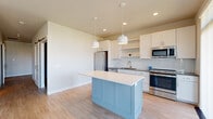 This single-floor unit presents a clean, contemporary design characterized by light hardwood-style flooring, neutral gray-toned walls, and white trim that runs consistently throughout. The kitchen serves as a central highlight, offering white shaker-style cabinetry, a striking herringbone-patterned tile backsplash, dark countertops, and a full suite of stainless steel appliances including a refrigerator, range, microwave, and dishwasher. A light blue center island with a crisp white countertop adds a distinctive color accent and provides additional workspace, illuminated by pendant lighting overhead. Recessed lighting complements the overall brightness of the kitchen area. A balcony access point off the kitchen introduces natural light and outdoor views of the surrounding cityscape. The bedrooms are generously proportioned, with one featuring large corner windows that invite abundant natural light and urban views, while another includes a bifold-door closet with wire shelving. Additional rooms offer clean, flexible spaces suited for various uses. A utility area with connections for laundry is also present. The unit maintains a cohesive, move-in-ready aesthetic throughout with consistent flooring and a neutral palette.