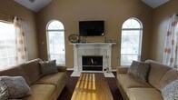 This single-story home showcases a thoughtfully designed living room that serves as its centerpiece. The space is defined by soaring vaulted ceilings with warm beige walls that lend an open, airy feel. Two striking arched windows with decorative fan transoms flank a white-mantled fireplace, flooding the room with natural light while offering views of the surrounding exterior. The fireplace itself serves as the focal point of the room, featuring a classic white surround and mantel accented with tasteful décor, with a wall-mounted flat-screen television positioned above. Warm-toned hardwood floors run throughout, complemented by a richly patterned area rug that anchors the seating arrangement. Comfortable tan leather sofas with decorative throw pillows face each other across a wooden coffee table, creating an inviting space for relaxation or entertaining. The combination of natural light from the arched windows, the warmth of the fireplace, and the rich wood flooring gives this living space a welcoming and refined ambiance that balances comfort with classic architectural detail.