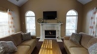 This single-story home showcases a thoughtfully designed living room that serves as its centerpiece. The space is defined by soaring vaulted ceilings with warm beige walls that lend an open, airy feel. Two striking arched windows with decorative fan transoms flank a white-mantled fireplace, flooding the room with natural light while offering views of the surrounding exterior. The fireplace itself serves as the focal point of the room, featuring a classic white surround and mantel accented with tasteful décor, with a wall-mounted flat-screen television positioned above. Warm-toned hardwood floors run throughout, complemented by a richly patterned area rug that anchors the seating arrangement. Comfortable tan leather sofas with decorative throw pillows face each other across a wooden coffee table, creating an inviting space for relaxation or entertaining. The combination of natural light from the arched windows, the warmth of the fireplace, and the rich wood flooring gives this living space a welcoming and refined ambiance that balances comfort with classic architectural detail.