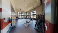 This modern fitness center is thoughtfully designed to support a full range of workout routines in a bright, open environment. The facility is divided into two distinct areas: a comprehensive main gym floor and a dedicated spin studio, each offering a focused and functional workout experience.

The main gym floor features soaring double-height ceilings and expansive multi-pane windows that line the walls, bathing the space in abundant natural light and offering open outdoor views. The floor is finished with durable rubber and hard-surface flooring, and the area is equipped with a robust selection of cardio machines—including treadmills and ellipticals—as well as strength-training stations, a full dumbbell rack, adjustable benches, and stability balls. Ceiling fans provide additional air circulation throughout the space, and wall-mounted televisions enhance the workout experience. Red accent stripes along the walls add a bold, energetic visual element.

The spin studio is a sleek, dedicated cycling space outfitted with multiple stationary bikes and floor-to-ceiling mirrors that create a sense of depth and openness. Warm wood-paneled accents, vibrant wall art, and striped flooring give the studio a polished, boutique aesthetic. Large windows continue the theme of natural light and outdoor connectivity found throughout the facility.