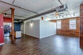 Located in the heart of Kansas City's downtown at 201 Main St, this single-floor loft-style space blends industrial character with modern comfort. The open living area makes a strong architectural statement with exposed brick walls, large arched windows that invite natural light, and oversized metallic ductwork running across the ceiling — all hallmarks of Kansas City's historic urban loft aesthetic. Rich, dark hardwood-style flooring flows throughout, complementing the bold accent walls in deep red and warm orange tones. Ceiling fans and track lighting provide both function and style overhead.

The kitchen is well-appointed with light maple cabinetry, dark granite-style countertops, stainless steel appliances including a range, microwave, and dishwasher, and a central island that offers additional prep and storage space. Bold red walls add a striking visual contrast to the warm wood tones of the cabinetry.

The bathroom features a granite-topped vanity with an undermount sink, a large mirror, and a full tub-shower combination, finished with cork-style flooring for added warmth. Structural wooden columns and exposed ceiling elements throughout the space reinforce the authentic industrial-loft character that defines this unique Kansas City address.