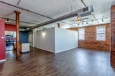 Located in the heart of Kansas City's downtown at 201 Main St, this single-floor loft-style space blends industrial character with modern comfort. The open living area makes a strong architectural statement with exposed brick walls, large arched windows that invite natural light, and oversized metallic ductwork running across the ceiling — all hallmarks of Kansas City's historic urban loft aesthetic. Rich, dark hardwood-style flooring flows throughout, complementing the bold accent walls in deep red and warm orange tones. Ceiling fans and track lighting provide both function and style overhead.

The kitchen is well-appointed with light maple cabinetry, dark granite-style countertops, stainless steel appliances including a range, microwave, and dishwasher, and a central island that offers additional prep and storage space. Bold red walls add a striking visual contrast to the warm wood tones of the cabinetry.

The bathroom features a granite-topped vanity with an undermount sink, a large mirror, and a full tub-shower combination, finished with cork-style flooring for added warmth. Structural wooden columns and exposed ceiling elements throughout the space reinforce the authentic industrial-loft character that defines this unique Kansas City address.