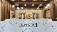 Located on Collins Road in Jacksonville, FL, the Tapestry clubhouse and leasing center is a single-floor facility that delivers a polished, contemporary aesthetic throughout its well-appointed spaces. Guests are greeted at a striking marble-clad reception desk beneath industrial-style pendant lighting, setting an upscale tone from the moment of arrival. The open lounge area beyond features soaring ceilings, a warm wood-paneled accent wall with a wall-mounted TV, and a chandelier centerpiece, all complemented by neutral tones accented with teal furnishings and curated wall art. A dedicated business and co-working nook offers round tables, teal velvet seating, pendant lighting, and built-in workstations with computers, ideal for residents needing a productive workspace. The leasing office area is bright and airy, with large windows, marble-finished countertops, and elegant drapery that allow ample natural light. A well-equipped kitchen and gathering space showcases a textured stone backsplash wall, expansive windows overlooking the exterior, pendant globe lighting, and a large central island — perfect for community events. Throughout, large-format tile flooring, contemporary artwork, and a cohesive coastal-modern palette create a welcoming and functional community hub.