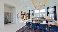 This contemporary single-floor amenity space makes an immediate impression upon entry, where oversized glass double doors open onto a spacious, light-filled interior anchored by a bold, patterned area rug and soaring double-height ceilings. The grand main lounge serves as the heart of the space, featuring a striking two-story wall of windows that floods the interior with natural light. A dramatic linear chandelier hangs above, complementing a warm wood built-in shelving unit that displays curated décor. The open-concept layout seamlessly connects a formal dining area with sleek dark chairs, a plush seating lounge with a fireplace, and a stylish reception desk accented by sculptural metallic wall art. Curated artwork, including bold abstract canvases and vibrant floral paintings, is displayed throughout, enhancing the sophisticated ambiance. A dedicated game room features a full-size billiard table beneath pendant lights, exposed beam ceilings, and generous windows overlooking the outdoors. A secondary lounge nook offers comfortable barrel chairs, a mounted flat-screen television, and direct access to the exterior. The overall design blends clean white walls, textured carpeting, and contemporary furnishings to create a polished, welcoming, and multifunctional community space.