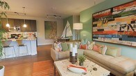 This single-floor space showcases a thoughtfully designed interior with a coastal-inspired aesthetic. The living area serves as the centerpiece, featuring rich hardwood flooring that extends throughout and adds warmth to the overall ambiance. A bold sage green accent wall provides a striking backdrop, complemented by neutral beige tones on the surrounding walls, creating a balanced and inviting color palette.

The living space is furnished with a generously sized sectional sofa adorned with an array of colorful throw pillows in coral, teal, and botanical prints, reinforcing the coastal theme. A stone-topped coffee table anchors the seating area and displays decorative accents, including candle holders and a model sailboat that adds a nautical charm. Large-scale artwork on multiple walls contributes character and visual depth to the room.

Track lighting along the ceiling provides adjustable illumination, enhancing the art and furnishings below. The layout flows seamlessly into an adjacent kitchen, visible in the background, which features dark cabinetry and a light countertop. The open-concept design promotes a sense of spaciousness and connectivity throughout the living space.