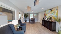 This single-floor space offers an open, well-connected layout that combines a living area and dining space in a cohesive, functional design. Light hardwood-style flooring runs throughout, lending warmth and continuity to the interior. Recessed lighting in the ceiling provides consistent illumination, complemented by natural light from windows along the far wall, which brighten the space considerably.

The living area features upholstered seating with nailhead trim detail, accompanied by side tables and a table lamp, creating an inviting, comfortable setting. A tall potted fiddle-leaf fig tree adds a natural, organic element to the room. Crown molding along the ceiling adds a refined architectural touch, while neutral gray walls serve as a versatile backdrop.

The dining area is positioned toward the rear of the space, furnished with a wooden table and matching chairs. Curtain-framed windows allow additional natural light into this zone. A dark wood sideboard along one wall offers storage and display space, topped with a coffee station and accented by a vibrant, colorful piece of artwork above. The overall ambiance is modern, clean, and welcoming.