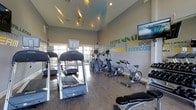 This single-floor fitness center is designed to provide a comprehensive and motivating workout environment. The space is outfitted with a strong selection of cardio equipment, including treadmills, stationary bikes, elliptical machines, and a rowing machine, catering to a variety of fitness preferences and training styles. A wall-mounted flat-screen television adds entertainment value for users during their sessions. The interior is brightened by large windows that line one wall, allowing generous natural light to flood the space and offer views of the outdoors, creating an open and energizing atmosphere. Soaring vaulted ceilings further enhance the sense of spaciousness, preventing the room from feeling cramped despite its full complement of equipment. The flooring is finished in warm hardwood tones, lending a clean and polished aesthetic. A bold, colorful typographic mural spans one wall, featuring motivational fitness-themed words such as "Determination," "Exercise," "Sport," and "Fitness," adding a dynamic and energetic visual element to the space. Recessed lighting supplements the natural light, ensuring the room remains well-illuminated throughout the day. Overall, the fitness center strikes a balance between functionality and an uplifting, purposeful design.