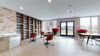Located at 150 Main St in Hackensack, New Jersey, this single-floor commercial space combines contemporary design with warm industrial aesthetics to create a highly functional and inviting environment. The main lounge area is defined by striking reclaimed wood shelving that lines an entire wall, providing both visual texture and display storage. Light wood-look flooring runs throughout, complementing the space's bright, airy atmosphere enhanced by recessed lighting and large windows that invite natural light and offer views of the surrounding neighborhood. Bistro-style tables paired with bold rust-orange chairs furnish the lounge, creating a casual yet polished setting ideal for informal meetings or collaborative work. A built-in kitchenette station is conveniently integrated into the lounge area. The meeting room features an exposed brick accent wall as its centerpiece, anchored by a wall-mounted display screen and solid wood live-edge conference tables with black chairs — a blend of rustic and modern sensibilities. Black-framed glass partition walls define and separate spaces while maintaining an open, connected feel throughout. A sleek, dark-paneled entryway corridor with glass walls on either side provides a refined transition into the interior. French doors in the lounge open directly to an outdoor area, further extending the usable space and bringing in additional natural light.