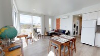This thoughtfully designed single-floor space offers an open-plan layout that seamlessly blends the living and dining areas into one cohesive, welcoming environment. Light hardwood flooring runs throughout, complementing the crisp white walls and recessed lighting that keep the interior bright and airy.

The living area is anchored by a dark leather sectional sofa adorned with accent pillows, paired with a classic wingback chair and a decorative textile wall hanging that adds a warm, artistic focal point. A wall-mounted mini-split system provides efficient climate control, while large floor-to-ceiling sliding glass doors and a tall vertical window flood the space with abundant natural light and frame expansive city and mountain views beyond.

The dining area features a handsome mid-century modern wooden table surrounded by matching chairs with upholstered seats, ideal for entertaining. The open sightlines between both zones create a generous sense of space and connectivity.

Tucked conveniently into an alcove just off the main living area is a stacked washer and dryer unit, offering in-unit laundry without interrupting the flow of the open floor plan. The overall ambiance is modern yet warm, blending clean architectural lines with carefully chosen furnishings to create a comfortable and functional living environment.
