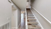 This two-story home offers a thoughtfully organized layout spread across two floors, with the main living areas on the ground floor and the bedrooms situated above. The welcoming lower level features an open-plan living, dining, and kitchen area bathed in natural light from large sliding glass doors adorned with vertical blinds. The living room is anchored by a tile-surround fireplace and ceiling fan, creating a comfortable focal point. The kitchen is well-equipped with white cabinetry, granite-style countertops, black appliances, and a charming bay window nook that brings in additional natural light. Light-toned laminate flooring flows throughout both levels, complementing the neutral gray and white color palette. Carpeted stairs with a white railing lead to the upper floor, where two bedrooms offer a private retreat. The spacious primary bedroom features its own ceiling fan, a natural light window with vertical blinds, and direct access to a vanity area. Each of the three bathrooms is cleanly finished, with one featuring a tiled tub-shower combo with warm beige tiles, and another with a white-tiled walk-in shower. Textured ceilings throughout add a classic residential character to the home.