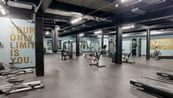 Located at 21 Erie Blvd in Albany, New York, this fitness facility offers a single-floor exercise space with an industrial-inspired aesthetic and a motivating atmosphere. The open floor plan is defined by exposed concrete ceilings, bold black structural columns, and dark overhead ductwork, all contrasted against light blue walls that lend an energetic yet focused ambiance. Durable rubber flooring tiles cover the entire workout area, providing comfort and safety underfoot.

The space is generously sized, accommodating a wide variety of training modalities. A dedicated cardio zone features treadmills, stationary bikes including air bikes, and rowing machines. The strength training area is well-equipped with a full dumbbell rack, weight benches, cable machines, and functional training equipment such as a sled, heavy punching bags, and a tire for cross-training exercises. Large wall mirrors throughout enhance the sense of space and allow for proper form monitoring during workouts.

Bold gold motivational phrases — including "Everyday is a Good Day to Work Out," "Never Give Up," and "No Time for Excuses, Only Results" — are emblazoned across the walls, reinforcing an encouraging training environment. Bright linear lighting ensures the space is well-illuminated throughout. The overall design balances raw, industrial character with a practical and energizing workout environment suited for a broad range of fitness routines.