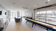 Located at 6137 Telluride Way in Denver, CO, this contemporary single-floor amenity space is thoughtfully designed for both socializing and relaxation. Light hardwood flooring runs throughout, complementing a clean, modern aesthetic defined by neutral tones and abundant natural light streaming in through floor-to-ceiling windows and glass-paneled walls.

A standout feature is the full-length shuffleboard table positioned at the center of the space, serving as a natural focal point for the game and entertainment area. Adjacent to it, a glass overhead garage door opens seamlessly to an outdoor terrace, blurring the boundary between indoor and outdoor living.

The kitchenette area is anchored by a striking textured stone accent wall in cool blue-grey tones, housing built-in cabinetry, a sink, appliances, and a coffee station. Mounted flat-screen TVs add a media element to this zone. Nearby, a large oval bar-height table with upholstered seating provides a gathering spot for dining or socializing.

A series of high-top bistro tables paired with sleek bar stools line one wall, accented by a vibrant 3x3 gallery wall of colorful pop-art prints, adding a playful, curated energy to the space. Additional small seating clusters with modern chairs are arranged throughout for more intimate conversation. Glass-walled rooms visible from the main floor suggest access to private meeting or fitness spaces.