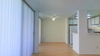 This single-floor space presents a clean, functional layout centered around an open living area. The room features light hardwood-style flooring that extends throughout, lending warmth and continuity to the interior. A ceiling fan is centrally mounted, providing both air circulation and ambient lighting. The textured popcorn ceiling adds a classic residential character overhead. Large vertical blinds line one wall, likely concealing a sliding glass door or wide window, allowing for adjustable natural light and privacy. The neutral beige walls create a bright, understated backdrop throughout. A structural column is visible near the blinds, defining the space without fully enclosing it. Toward the rear of the living area, an opening leads into a kitchen zone, where upper cabinetry and a refrigerator are partially visible, suggesting a practical and accessible layout. The transition between the living and kitchen areas is open, promoting a connected flow between the two spaces. Overall, the space maintains a simple, well-lit atmosphere with functional finishes suited for everyday living.