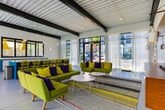 Located on Boulevard SE in Atlanta's dynamic Grant Park neighborhood, this single-floor amenity space showcases a bold, contemporary design that blends industrial architecture with vibrant interior styling. Exposed metal deck ceilings with dark structural steel beams lend an industrial-chic character throughout, while polished concrete floors provide a clean, modern foundation underfoot.

The lounge area is a standout feature, anchored by a striking accent wall of warm horizontal wood paneling housing a large flat-screen television and built-in cabinetry. Chartreuse and olive-toned sofas and chairs, accented with bold purple and yellow cushions, are arranged around a colorful striped area rug and sleek white side tables, creating an inviting and energetic gathering space. Floor-to-ceiling sheer curtains filter abundant natural light streaming in from large windows, which also offer views of the surrounding urban streetscape.

Adjacent to the lounge, a generously sized kitchen and social area features a prominent teal island with integrated sink, lined with a row of bright yellow metal bar stools. White cabinetry lines the perimeter wall beneath a display window, and globe pendant lights hang overhead. Double glass entry doors flood the space with additional daylight and connect it visually to the outdoors, reinforcing an open and welcoming atmosphere throughout.