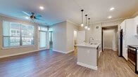 This single-floor residence is designed with a clean, contemporary aesthetic throughout. The open-concept living and kitchen area serves as the heart of the unit, featuring warm-toned wood-look flooring, crown molding, and recessed lighting complemented by a ceiling fan. A central kitchen island with a granite countertop and undermount sink anchors the space, illuminated by a trio of pendant lights. The kitchen is fully equipped with white shaker-style cabinetry, stainless steel appliances including a refrigerator, range, and over-the-range microwave, and a classic subway tile backsplash. Natural light flows into the living area through large windows fitted with horizontal blinds, with a door providing exterior access. The bedroom offers plush carpeting, dual windows, a ceiling fan, and access to a spacious walk-in closet outfitted with wire shelving. The bathroom features wood-look flooring, a floating dual-sink vanity with granite countertops, two framed mirrors, and black hardware accents. A dedicated laundry area accommodates a full-size side-by-side washer and dryer, completing the functional layout of this well-appointed space.