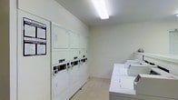 Located at 715 Moon Road in Columbus, Ohio, this single-floor shared laundry facility is designed to serve residents with a practical and functional layout. The room features bright overhead fluorescent lighting that illuminates the clean, white interior, creating a well-lit environment for everyday use. The walls are finished in a simple white painted brick, contributing to the room's tidy and utilitarian aesthetic. The floor is covered in light-colored tile, making maintenance and cleanliness straightforward. Along one wall, stacked coin-operated dryers are neatly arranged, providing ample drying capacity. Opposite, a row of top-load washing machines lines the adjacent wall, each equipped with individual compartments, offering users convenient access and efficient use of space. Posted instructions are clearly visible near the entrance, ensuring ease of use for all residents. The space is thoughtfully organized to maximize the number of machines available while maintaining comfortable circulation throughout the room. Overall, this laundry facility offers a clean, no-frills environment focused entirely on functionality and accessibility for the building's residents.