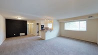 This single-floor residence offers an open-concept layout that seamlessly connects the living area to the kitchen. The spacious living room is finished with wall-to-wall carpet in a neutral tone, creating a comfortable and cohesive feel throughout. A large window along the far wall allows abundant natural light to fill the space, offering views of surrounding greenery.

The kitchen is positioned centrally, defined by a peninsula with an overhang that accommodates seating and serves as a natural divider between the cooking and living areas. White cabinetry lines the kitchen walls, complemented by a dark countertop and a track lighting system overhead that provides focused task lighting. A microwave and additional appliances are integrated into the kitchen setup. A window above the sink adds further brightness to the cooking area.

Adjacent to the kitchen, a stacked in-unit washer and dryer are conveniently tucked into a dedicated laundry space. A hallway visible in the background leads to additional rooms, suggesting a functional and well-organized floor plan. The overall ambiance is clean, bright, and practical.