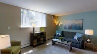 This single-floor residential space offers a comfortable and well-appointed living environment designed with everyday functionality in mind. The living room serves as the heart of the space, featuring wall-to-wall carpeting that adds warmth underfoot and contributes to the room's cozy atmosphere. A large window dressed with vertical blinds allows an abundance of natural light to fill the room, creating a bright and airy feel throughout the day. The walls are painted in complementary neutral and soft teal tones, lending the space a calm, modern character. A wall-mounted air conditioning unit ensures year-round climate comfort, while baseboard heating runs along the perimeter for additional warmth. The room is thoughtfully arranged with a plush sofa accented by decorative pillows, a sleek coffee table, and a distinctive multi-toned storage cabinet that doubles as a decorative focal point. Ambient lighting from a table lamp and a multi-armed floor lamp adds a layered, inviting glow to the space during the evening hours. A piece of abstract wall art in cool blue tones complements the overall color palette, tying the room's aesthetic together cohesively. The space strikes a balance between practical comfort and stylish design, making it a welcoming retreat.