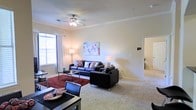 This single-floor space offers a thoughtfully designed interior that balances comfort and style. The living area serves as the heart of the home, featuring plush wall-to-wall carpeting that adds warmth underfoot. Sleek black leather seating arranged around a glass-topped coffee table creates an inviting focal point, while a bold area rug in deep red adds a striking contrast to the neutral palette. A ceiling fan with integrated lighting ensures the space remains comfortable throughout the year. Natural light streams in through a large window dressed with sheer white curtains, brightening the room and enhancing its airy feel. The walls are finished in a soft, neutral tone, providing a clean backdrop that allows the colorful abstract artwork to stand out as a decorative centerpiece. A dedicated dining area is visible at the edge of the living space, complete with a dark-toned table and coordinating chairs. A hallway leads to the remaining areas of the unit, suggesting a well-organized layout. The overall ambiance is modern and inviting, with a cohesive design scheme that makes the space feel both functional and stylish.