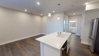 This single-floor space showcases a clean, contemporary design with a neutral color palette throughout. The open-concept kitchen and living area serves as the heart of the unit, anchored by a large center island with a white quartz countertop, an integrated sink, and elegant pendant lighting overhead. White shaker-style cabinetry lines the kitchen walls, complemented by stainless steel appliances including a refrigerator, range, and over-the-range microwave. Dark hardwood flooring extends throughout the main living and kitchen areas, adding warmth and contrast to the bright, white finishes. Recessed lighting provides ample illumination across the open space, while a transom window above the entry door allows additional natural light into the kitchen corridor. The bedroom offers a comfortable retreat, featuring plush carpet flooring, a ceiling fan with integrated lighting, and a window dressed with blinds that filters in natural light. The full bathroom is finished with a white vanity, quartz countertop, framed mirror, and a combination tub and shower, maintaining the cohesive modern aesthetic found throughout. The overall ambiance is fresh, bright, and move-in ready.