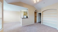 This single-story residence is designed with an open, functional layout that maximizes comfort and usability throughout. The living area serves as the heart of the home, showcasing soaring vaulted ceilings and a ceiling fan that enhance the sense of spaciousness. A standout architectural detail is the built-in arched shelving unit, offering both display and storage space. The living area connects seamlessly to the kitchen via a pass-through counter with a dark countertop, allowing for easy interaction between spaces. The kitchen is well-appointed with light wood cabinetry extending to the ceiling, a white appliance suite including a refrigerator, electric range, and microwave, and tile flooring that contrasts with the plush carpeting found throughout the rest of the home. The bedrooms are generously sized, each featuring carpet flooring, ceiling fans, and windows that allow natural light to fill the rooms. One bedroom includes direct access to an en-suite bathroom. Both bathrooms are finished with tile flooring, granite-style countertops, wood-toned vanity cabinetry, and tub-shower combinations with white tile surrounds. The overall color palette of warm beige tones creates a cohesive and neutral ambiance throughout the home.