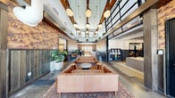 Located at 920 Loveland Dr in Stillwater, Oklahoma, this single-floor community clubhouse offers a generously sized, thoughtfully designed gathering space that blends industrial and boutique aesthetics. The centerpiece is a dramatic, double-height lobby featuring soaring ceilings with exposed wooden beams, a cluster of globe pendant lights, and an upper mezzanine level lined with lush greenery. The walls are adorned with distinctive toile wallpaper and gray shiplap wainscoting, giving the space a warm, eclectic character. Tufted leather sofas are arranged around a central coffee table atop a layered vintage rug, creating an inviting lounge atmosphere. A staffed reception desk anchors one end of the lobby, while a unique suspended globe chair adds a playful focal point near large glass doors that flood the space with natural light. Adjacent to the main lounge is a conference-style room featuring a long communal table, a patterned ceiling, and glass-panel partitions that offer both openness and privacy. The fitness center is well-equipped with a comprehensive selection of strength machines and cardio equipment, wall-to-wall mirrors, and ceiling fans, providing a fully functional workout environment. Polished concrete floors flow throughout, tying all areas together with a cohesive, contemporary feel.