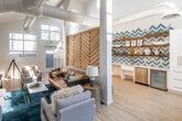 Located at 310 Jefferson St in Placentia, CA, this single-floor community space delivers a striking blend of contemporary design and relaxed, coastal-inspired style. The soaring vaulted ceilings amplify the sense of openness, while exposed white ductwork adds an industrial-chic character that complements the clean, bright interiors. Large floor-to-ceiling windows flood the space with natural light, offering views of the surrounding outdoor areas and keeping the atmosphere vibrant throughout the day. Light wood-tone flooring runs throughout, grounding the space with warmth and continuity. The main lounge area is anchored by a rich cognac leather sofa, coordinating gray seating, and a bold teal area rug, centered around a flat-screen TV mounted above a dark media console with rustic floating wood shelves. A large slatted wood chevron wall panel serves as a dramatic architectural focal point, adding texture and visual depth. A separate dining nook features a round stone-topped table with upholstered chairs beneath expansive windows. The kitchenette area stands out with a vivid blue-and-white chevron tile backsplash, open wood shelving, white shaker cabinetry with gold hardware, a beverage refrigerator, and a built-in microwave — creating a fully functional and visually arresting refreshment station.