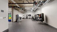 Located at 2108 Berkeley Way in Berkeley, California, this fitness center offers a generously sized, single-floor space designed to support a full range of workout routines. The facility features an industrial-inspired aesthetic, with exposed concrete ceilings, visible ductwork, and overhead pipe systems that give the space a modern, urban character. Bright linear LED lighting runs throughout, ensuring the entire floor is evenly and efficiently illuminated.

The gym is outfitted with an extensive selection of equipment to accommodate both cardio and strength training needs. Cardio machines include multiple treadmills, elliptical trainers, a stair climber, and a rowing machine, several of which are paired with wall-mounted flat-screen televisions for an engaging workout experience. The strength training area features a cable machine, a multi-functional rack loaded with medicine balls and dumbbells, weight benches, and a suspended heavy punching bag for boxing and conditioning. A mirrored wall section expands the visual depth of the space and supports form-focused training.

The flooring throughout is durable rubber gym tile in a speckled grey finish, ideal for high-impact use. The layout is thoughtfully organized to allow comfortable movement between zones. A water fountain is also available on-site. The overall ambiance is functional, clean, and purpose-built for fitness.