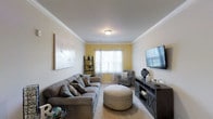 This single-floor residence offers a thoughtfully designed living space that balances comfort and style. The living room serves as the centerpiece of the home, showcasing warm yellow accent walls that complement crisp white crown molding and a clean white ceiling — a classic architectural detail that adds a refined character to the room. Plush wall-to-wall carpeting underfoot contributes to the cozy ambiance throughout. Natural light flows generously into the space through windows dressed with sheer white curtains, creating a bright and airy atmosphere. The room is furnished with a spacious gray sectional sofa accented with patterned throw pillows, a round upholstered ottoman as a central focal point, and a cushioned accent chair tucked into the corner. A dark wood media console anchors one wall, topped with a large flat-screen television, while a lean-to shelving unit near the window adds functional display space. A large abstract canvas on the wall adds an artistic touch without overwhelming the space. Overall, the residence presents a warm, welcoming environment with tasteful décor, cohesive tones, and practical layout choices that make it both livable and visually appealing.