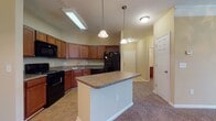 This single-floor residence offers a well-organized layout with a cohesive design highlighted by warm beige and tan wall tones, plush carpeted flooring in the living and bedroom areas, and tile flooring in the kitchen and bathroom. The open-concept kitchen serves as a central hub of the home, featuring rich medium-toned wood cabinetry, laminate countertops, a double stainless steel sink, and a full suite of black appliances including a range, over-the-range microwave, refrigerator, and dishwasher. A center island with pendant lighting above provides additional counter space and a natural transition into the adjoining living area. The spacious living room is finished with crown molding, a ceiling fan, and pendant light fixture, creating a comfortable and polished atmosphere. A separate dining area connects seamlessly to the kitchen. The bedrooms offer generous proportions, carpeted floors, and windows that admit natural light and neighborhood views. The bathroom includes a full tub-shower combination with tile surround, a wide vanity with rich wood cabinetry, laminate countertops, and a large mirror. The textured ceilings and white panel doors are consistent throughout, contributing to a clean and uniform interior aesthetic.
