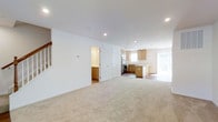This two-story home offers a well-designed open-concept main level that seamlessly connects the living area to the kitchen. The living space is finished with neutral wall-to-wall carpeting, creating a warm and inviting atmosphere, while recessed lighting throughout ensures consistent, bright illumination. Clean white walls and white trim contribute to a fresh, modern aesthetic.

The kitchen is outfitted with light wood cabinetry, stainless steel appliances, and a central island that provides additional workspace and storage. A window above the sink and a large window or sliding door near the dining area allow natural light to flow into the space. The kitchen transitions smoothly into the main living area, making the layout practical for everyday living and entertaining.

Adjacent to the living area, two interior doors lead to additional spaces, including what appears to be a utility or storage room with matching cabinetry. A staircase with a wooden banister along the left wall provides access to the upper floor. The overall design prioritizes a clean, functional layout with a neutral color palette that lends itself well to a variety of interior styles.