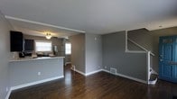 This two-story home offers a well-designed layout that balances functionality and style across both levels. The main floor showcases an open-concept living and kitchen area, unified by rich, dark hardwood flooring that runs throughout the space. The living area is generous in scale, featuring neutral gray walls, crisp white trim and baseboards, and a textured ceiling that adds subtle visual interest. Natural light pours in through multiple windows, creating a bright and welcoming atmosphere.

The kitchen flows seamlessly from the living area and is equipped with stainless steel appliances, including a refrigerator and gas range. A breakfast bar provides a natural dividing point between the kitchen and the main living space, making it ideal for casual dining or entertaining. A ceiling fan adds both practicality and a finishing touch to the kitchen area.

A staircase, framed by clean white railings, leads to the second floor, hinting at additional living space above. Floor vents are integrated neatly into the hardwood floors, and the overall design reflects a cohesive, modern aesthetic. The neutral gray-and-white color scheme throughout the main level creates a versatile backdrop that is both contemporary and inviting.