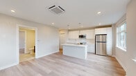 This single-floor unit showcases a clean, contemporary design throughout. The open-concept kitchen serves as the heart of the space, anchored by a large central island with a white quartz countertop and an undermount sink. Flat-panel white cabinetry lines the walls, complemented by stainless steel appliances including a refrigerator, range, and built-in microwave. Pendant lighting hangs above the island, while recessed ceiling lights provide additional illumination. Light-toned hardwood flooring flows seamlessly through the kitchen and into the adjoining living area, reinforcing the open, airy feel. Large windows in the living space and bedrooms invite natural light and frame outdoor views of the surrounding neighborhood. The bedrooms are well-proportioned, with one featuring dual windows and a ceiling-mounted HVAC unit, while another includes a sliding-door closet. A hallway with louvered storage doors connects the living areas to the private spaces. The bathroom is finished with large-format tile flooring, a floating dark-wood vanity with a white countertop, and a clean, minimalist aesthetic. Throughout the unit, neutral tones and modern finishes create a cohesive, move-in ready environment.