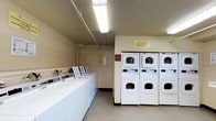 This laundry room provides a well-organized, functional space designed to accommodate multiple users efficiently. A row of top-loading washing machines lines one wall, offering ample capacity for laundry needs. Opposite, stacked dryer units are arranged side by side, maximizing the use of vertical space and providing several drying options simultaneously. The room features neutral-toned walls and a light-colored floor, contributing to a clean and utilitarian aesthetic. Overhead fluorescent lighting ensures the space is well-lit throughout. Exposed utility piping runs along the upper portion of the walls, reflecting the room's practical design focus. A utility panel is mounted on the wall, and informational signage is posted for users. The layout is straightforward, with clear circulation space between the machines, making it easy to move around and manage laundry tasks. Overall, the space is efficiently arranged to serve as a reliable shared laundry facility.