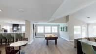 This single-floor amenity space is thoughtfully designed to offer a variety of social and recreational functions within a cohesive, modern aesthetic. The layout flows between distinct zones, each serving a unique purpose while sharing a consistent design language of neutral tones, clean lines, and patterned carpet flooring throughout.

The game room is a standout feature, centered around a full-size pool table with a warm caramel felt surface. Multiple windows allow ample natural light to fill the space, and a trio of framed green landscape prints adds a pop of color to the walls. A wall-mounted flat-screen television is also present for additional entertainment.

Adjacent to the game room, the lounge area provides a comfortable gathering spot furnished with plush seating, including a large sectional sofa and dark leather armchairs arranged around a round coffee table. A bold yellow abstract canvas anchors the room visually, while a row of windows along one wall invites natural light and outdoor views.

The kitchen is well-appointed with dark espresso cabinetry, white quartz countertops, a double stainless steel sink, pendant lighting, and a textured mosaic tile backsplash. Light-toned tile flooring, a coffee station, and ample counter space make it ideal for hosting and catering events.