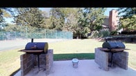 This outdoor recreational space is thoughtfully designed to accommodate a variety of leisure and social activities. The area features a covered pavilion supported by white structural columns, providing a shaded retreat with a concrete floor ideal for gatherings. Beneath the pavilion, a vibrant red round picnic table offers a comfortable spot for outdoor dining and socializing.

Adjacent to the pavilion, a dedicated grilling area is constructed with sturdy brick masonry bases supporting multiple barrel-style grills, making it well-suited for outdoor cooking and entertaining. The grilling stations are oriented to face a generous open lawn area, providing an inviting view while in use.

The expansive green lawn serves as a central feature of the space, bordered by a chain-link fence with privacy screening that encloses a tennis court — a notable amenity for active recreation. Mature, full-canopy trees line the perimeter, offering natural shade and a lush, park-like atmosphere throughout the grounds.

A concrete walkway connects the pavilion area to the broader grounds, with landscaped steps leading to additional outdoor amenities, including what appears to be a pool area visible in the background. Well-maintained shrubbery and green landscaping throughout the space contribute to a clean, organized, and welcoming outdoor environment suitable for community use.