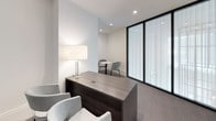 This single-floor office space is designed with a refined, contemporary aesthetic that balances functionality with sophistication. The layout includes a primary workspace anchored by a dark wood-toned desk with built-in storage drawers, accompanied by plush upholstered seating in neutral tones. A dedicated meeting nook features a round white bistro table paired with two blue accent chairs, set against floor-to-ceiling glass partitions fitted with sheer white curtains that diffuse natural light while maintaining privacy.

Recessed ceiling lighting throughout ensures consistent, even illumination across all areas. The space is finished with patterned grey carpeting that adds subtle texture underfoot, while crisp white walls with substantial baseboards lend a clean, polished character. Curated framed artwork depicting lush botanical scenes adds warmth and visual interest to the otherwise understated palette.

Entry is provided through elegant dark wood barn-style doors with mirrored glass panels, offering both style and practicality. A table lamp with a warm-toned shade contributes ambient lighting to the main workspace. Overall, the space conveys a calm, professional atmosphere well-suited for private office use or client-facing consultations.