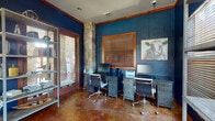 This well-appointed single-floor space blends rustic charm with modern functionality, creating an inviting environment suited for work and collaboration. The interior is defined by rich navy blue walls accented with warm wood crown molding and door framing, polished stained concrete floors, and a distinctive natural stone column that serves as a standout architectural focal point.

The workspace area is equipped with dual computer workstations, ergonomic rolling chairs, and a multi-drawer desk unit, providing ample setup for productivity. A metal open-shelf bookcase displays books and decorative objects, adding character alongside a rustic cow-themed wall painting that reinforces the space's eclectic aesthetic. Wood-slat blinds on the windows allow for adjustable natural light throughout.

A comfortable lounge area features two leather armchairs with accent pillows arranged around a round coffee table, offering a relaxed setting for casual meetings or reading. French doors with wood framing open to an outdoor area, flooding the space with natural light and providing a visual connection to the exterior. An additional seating nook near the entry holds a single leather armchair, creating a welcoming transition point. Overall, the space balances a professional atmosphere with warm, distinctive design details.