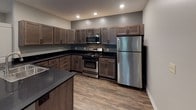 This single-floor residence offers a thoughtfully designed open-concept layout that seamlessly connects the living area and kitchen. The living room is spacious and welcoming, featuring rich wood-look plank flooring, neutral gray walls, and a standout accent wall clad in reclaimed-style wood planking that frames a built-in fireplace. A glass exterior door floods the space with natural light and provides access to the outdoors.

The kitchen is a functional and stylish centerpiece, outfitted with dark-stained wood cabinetry, sleek black countertops, and a full suite of stainless steel appliances including a gas range, built-in microwave, and refrigerator. Recessed lighting illuminates the space, while wood-look flooring ties the kitchen seamlessly to the living area. A kitchen island with a sink adds practical workspace and defines the transition between the two areas.

The bedrooms are finished with plush carpeting and neutral-toned walls, offering a comfortable and quiet retreat. Each room benefits from natural light via window blinds. The bathroom features a dark wood vanity with a black countertop and undermount sink, a tiled tub-and-shower combination, and neutral tile flooring, creating a clean and contemporary aesthetic throughout.