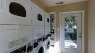 This single-story laundry facility is designed for shared use, offering a practical and functional layout. The room houses multiple stacked washer and dryer units arranged along the walls, maximizing the available floor space while accommodating several loads simultaneously. The machines are front-loading models, providing efficient and accessible operation. The interior features neutral beige-toned walls and light flooring, contributing to a clean and well-maintained atmosphere. Overhead ventilation ensures adequate airflow throughout the space. A glass-paneled door provides direct access to the outdoors, allowing natural light to filter into the room and offering views of the surrounding tree-lined exterior. A posted notice on the door reminds users to keep the door closed at all times, reflecting the facility's attention to security and upkeep. The overall design prioritizes utility and ease of use, making it a straightforward and convenient space for laundry needs.