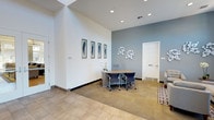 This single-floor leasing office offers a polished and professional environment designed to welcome prospective residents. The space features soaring ceilings that amplify the sense of openness, complemented by a clean, contemporary aesthetic throughout. The main reception and consultation area combines warm neutral flooring — transitioning from tile at the entrance to carpet in the workspace — with cool-toned blue-grey accent walls that add a calm, inviting atmosphere. Recessed lighting illuminates the space evenly, highlighting thoughtful decorative touches including a series of framed feather artwork and modern sculptural wall décor. A streamlined wooden consultation desk with ergonomic rolling chairs serves as the central workstation, providing a comfortable setting for one-on-one meetings with prospective residents. A fresh orchid plant on the desk adds a touch of natural elegance. A private door leads to an additional room, suggesting dedicated workspace or a private office for staff. The overall design balances functionality with a refined, contemporary style, creating a professional yet approachable environment suitable for leasing consultations and community inquiries.
