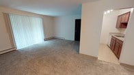 This single-floor unit offers a straightforward, functional layout with a neutral color palette throughout. The main living area is generously sized, featuring wall-to-wall carpet in a warm beige tone and a large sliding glass door fitted with vertical blinds, allowing abundant natural light and providing access to an outdoor balcony area with surrounding greenery. A baseboard heating system runs along the perimeter walls, and a wall-mounted air conditioning unit ensures year-round comfort.

The bedroom is a well-proportioned, carpeted room with a window overlooking the outdoors, a baseboard heater, and a closet. The textured ceilings are consistent throughout the unit, adding a classic residential character.

The kitchen is compact yet practical, featuring warm-toned wood cabinetry, a speckled laminate countertop, a stainless steel sink, and a white range. Light tile flooring distinguishes the kitchen from the carpeted rooms.

The bathroom includes a full tub and shower combination enclosed by clean white square tiles with chrome fixtures, maintaining a simple and functional aesthetic throughout the unit.